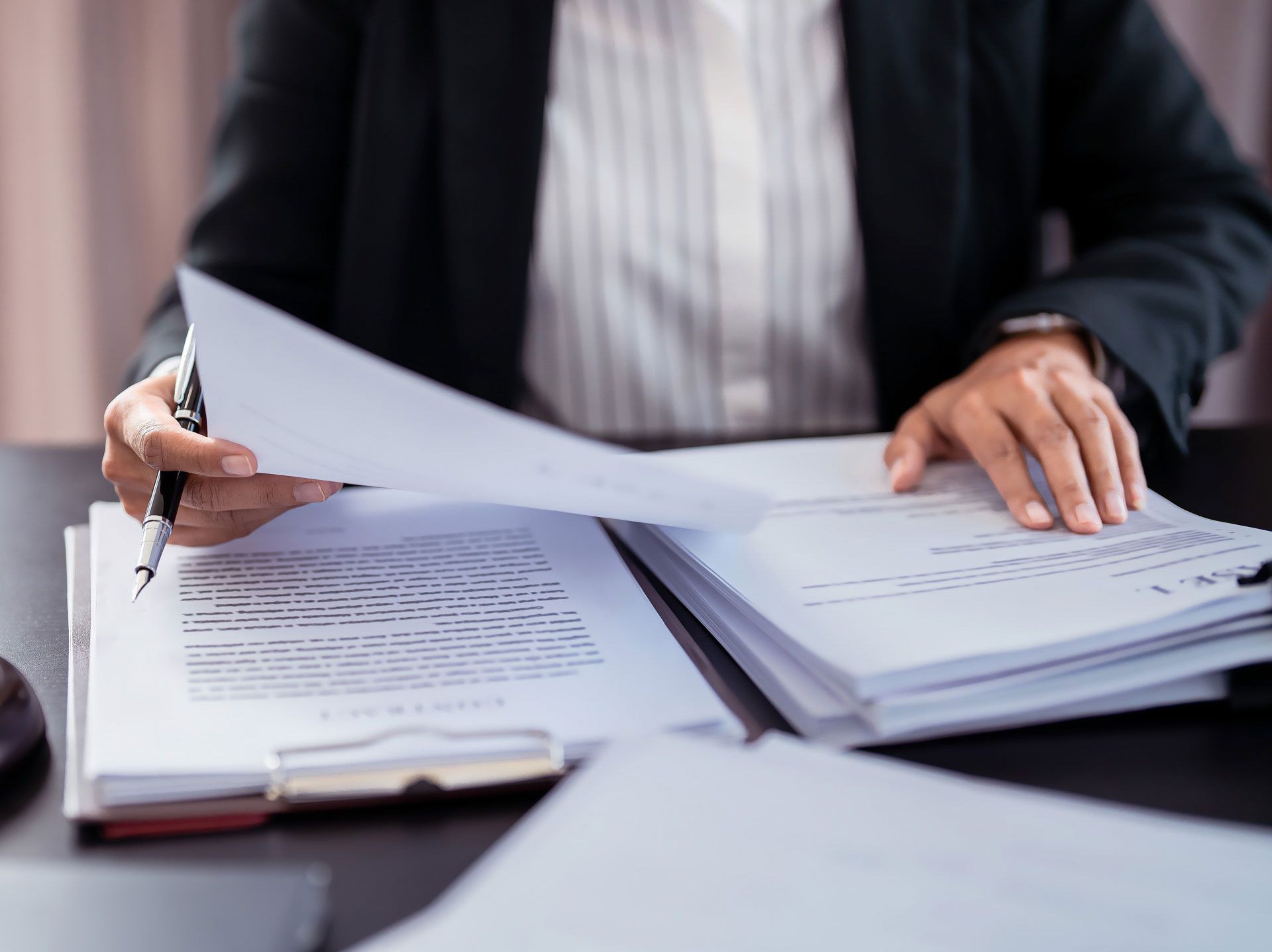Person in a blazer reviewing documents at a desk with a pen in hand.