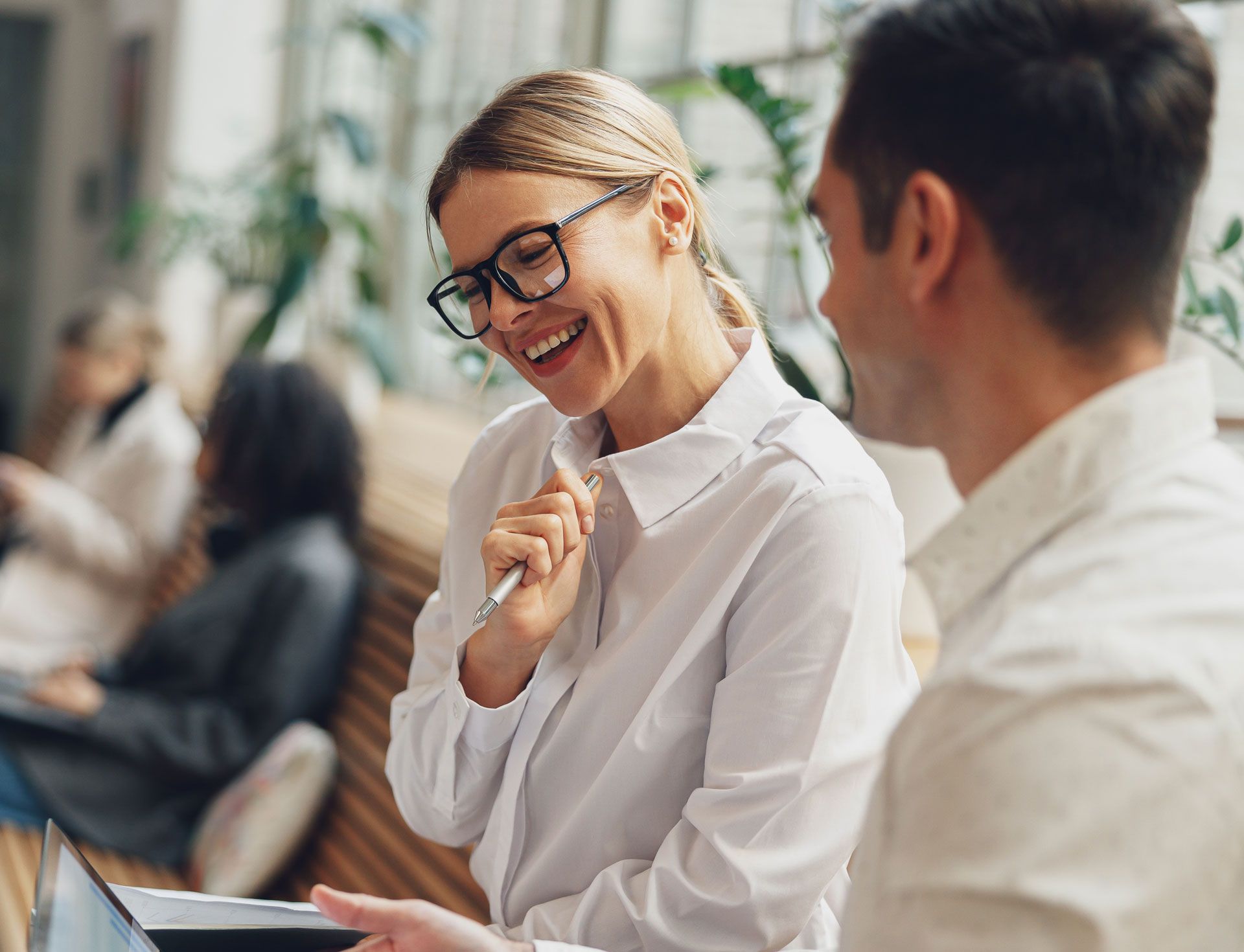 Woman with glasses laughs, pointing at a laptop screen with a man. They are in an office setting.