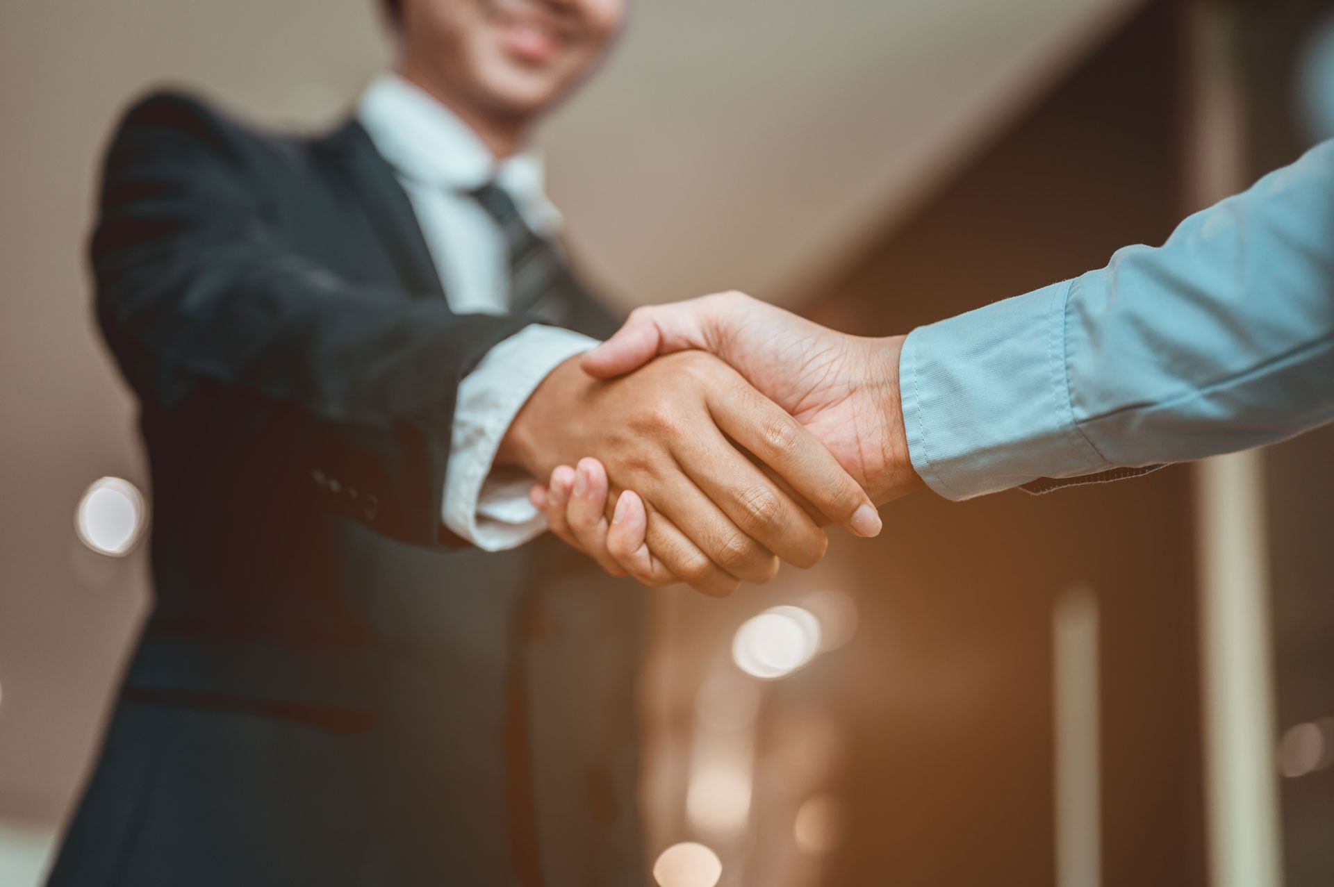 Two businessmen shaking hands, wearing suits, indoors, blurred background.