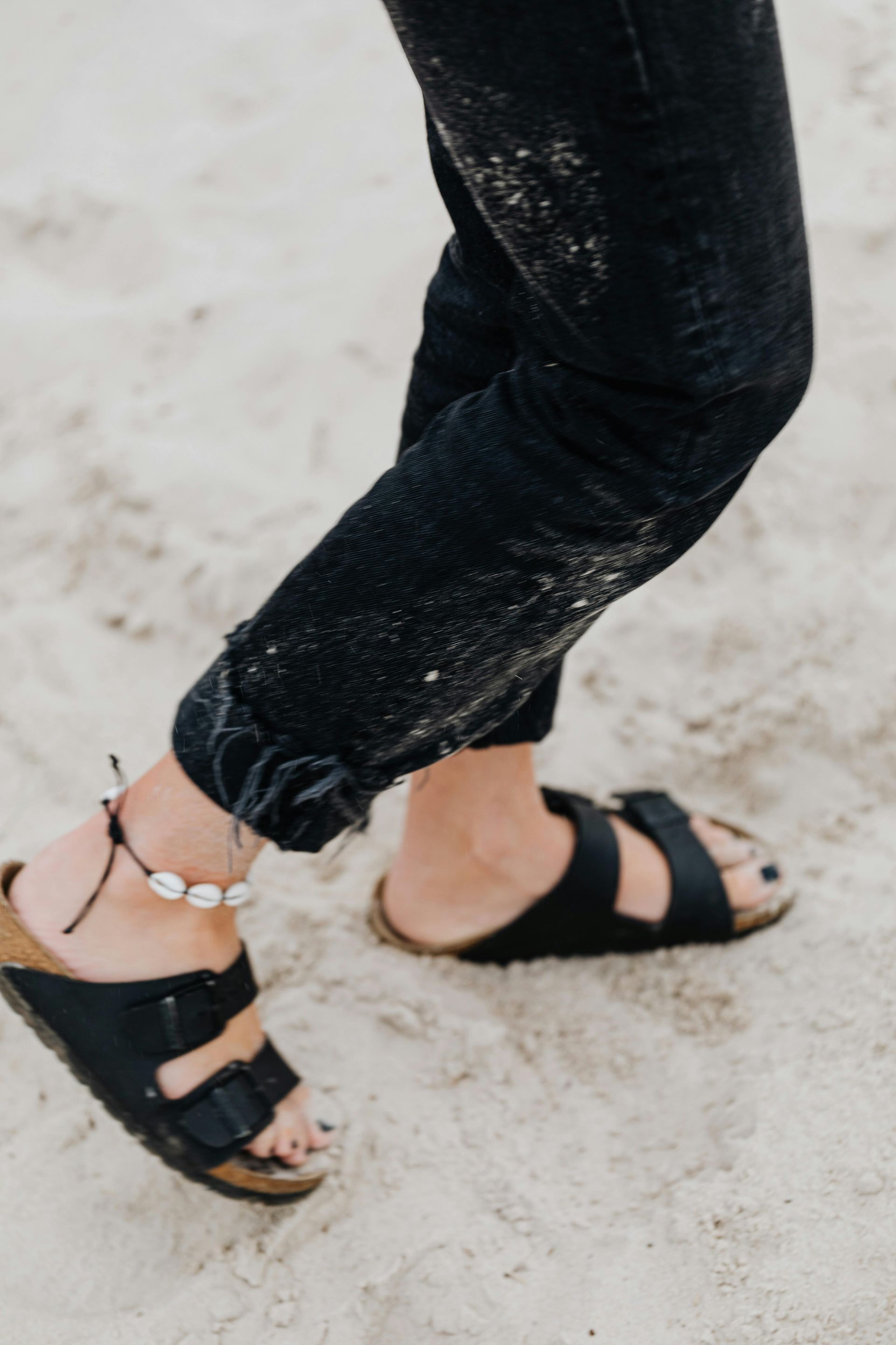cropped photo of woman's legs and feet, wearing black heans and crock black sandals with shell anklet. walking on the sand.