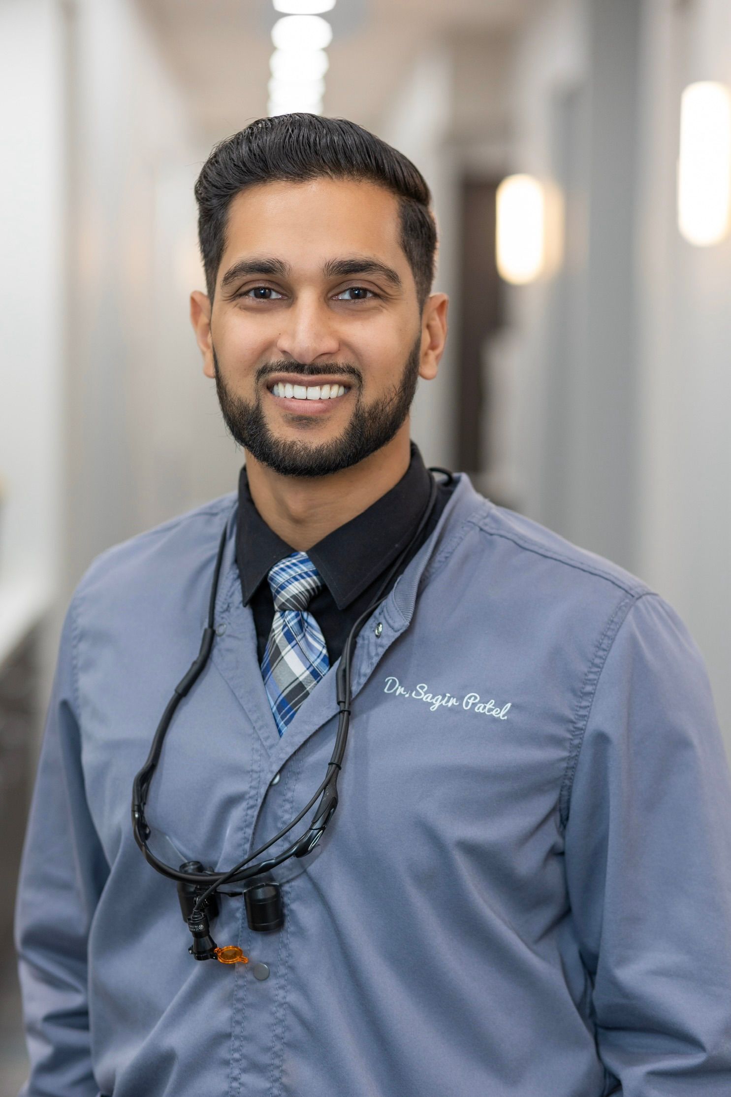 Smiling man in dental scrubs and tie, wearing dental loupes, in a hallway.