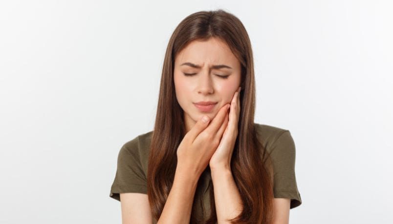 Woman with long brown hair, eyes closed, holding her cheek, likely experiencing tooth pain.