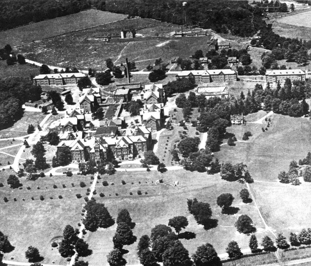 An aerial view of the Ontario Hospital School, Orillia as it appeared in the 1930s. Closest to the bottom of this image is the Administration Building, flanked on either side by Cottages K (left) and A (right). To the left of Cottage K is the newly constructed Hospital/Infirmary, which was used an admissions ward until the construction of the Isolation Ward in the mid-1950s. Behind the administration building is the Recreation Hall/Gymnasium and running back from this building is a long structure which contained facilities such as the institution kitchens and workshops. Between this structure and Memorial Avenue is the second institution Power House. On the left side of this long structure are Cottages L and M and on the right side are Cottages B and C. Running parallel to Memorial Avenue are three cottages constructed during S.J.W. Horne’s tenure as Superintendent. From left to right, these are Cottages O and D and the Nurses’ Residence. On the opposite side of the road is the institution farm. Three small farm houses face the road and beyond these, from left to right, are the cow barn, piggery and water tower, horse barn and blacksmith shop. To the right of the blacksmith shop is the institution cemetery. Image from the Regional Centre Records.)