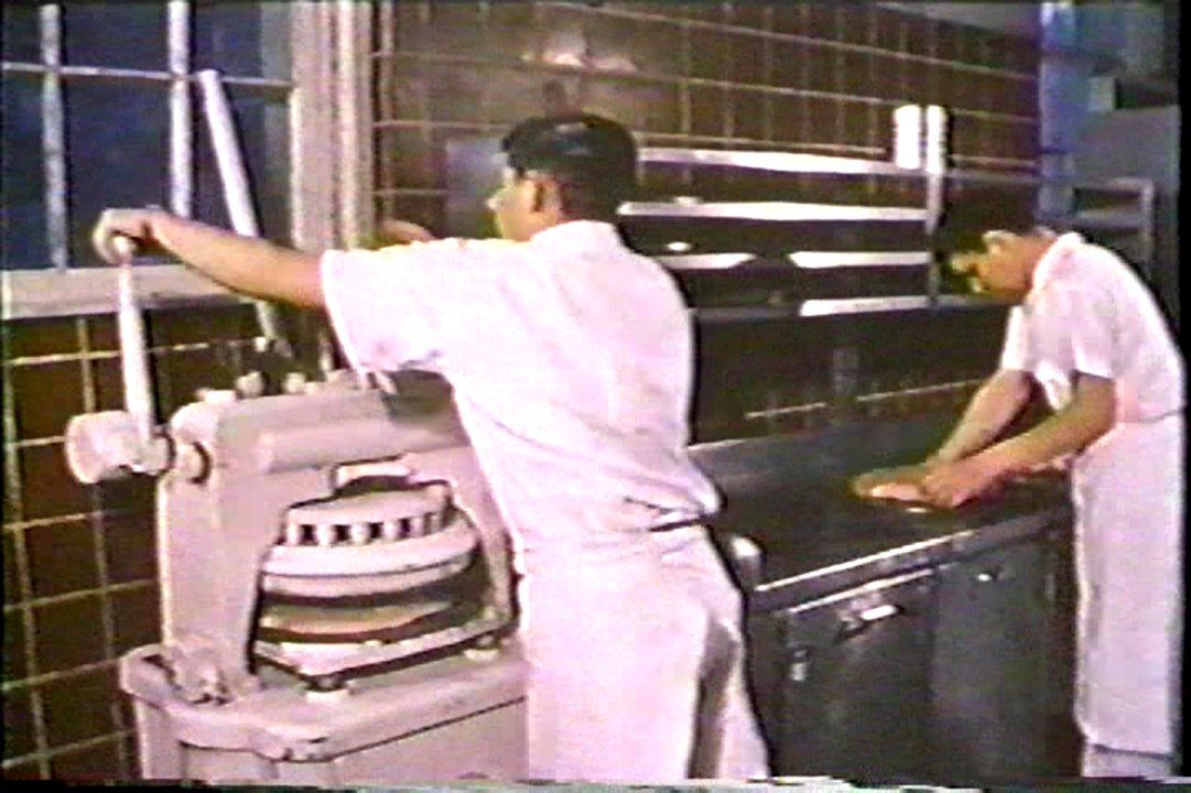 Two male residents working in the institution bakery in 1960. One is operating a press that molds dough into dinner roles and the other appears to be kneading dough on a metal counter. Image from the government propaganda film One On Every Street.