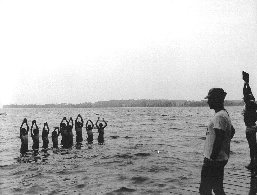 This photograph shows nine boys swimming at one of the Huronia Regional Centre beaches. They are standing in a row on the left side of the image with their hands over their heads and water up to their waists. On the right side of the image, two male staff members stand on a wooden dock. The staff members are wearing swim trunks and t-shirts. One is holding a clipboard over his head. Image from the Regional Centre Records.