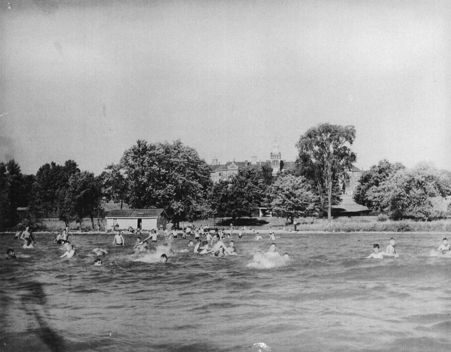 Boys swimming at one of the institution’s beaches, as seen from the lake. On the shoreline beyond is a small wooden building, probably a changing room or washroom. Beyond this is the Canadian National Railway line and a grouping of trees. In the distance, above the trees, the Administration Building and Cottages A and K can be seen. Image from the Regional Centre Records.