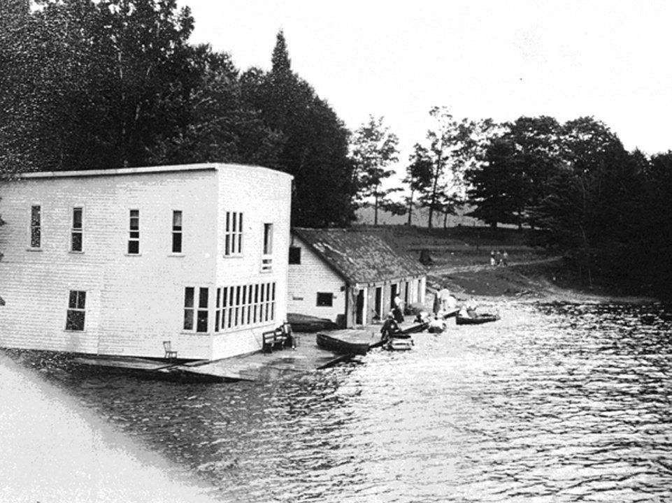 A photograph of the boathouse at the Huronia Regional Centre taken in the 1920s. The image shows two large wooden buildings on a wooden dock. One is a two storey square building that is painted white and has numerous windows. The other is a large one storey building with five large doors for bringing boats in and out. Several boats are in the water or on the dock. This boathouse was demolished at an unknown point and replaced with a smaller structure that still stands today. It is unclear which boathouse Hilda Sturman was killed in. Image from the Regional Centre Records.