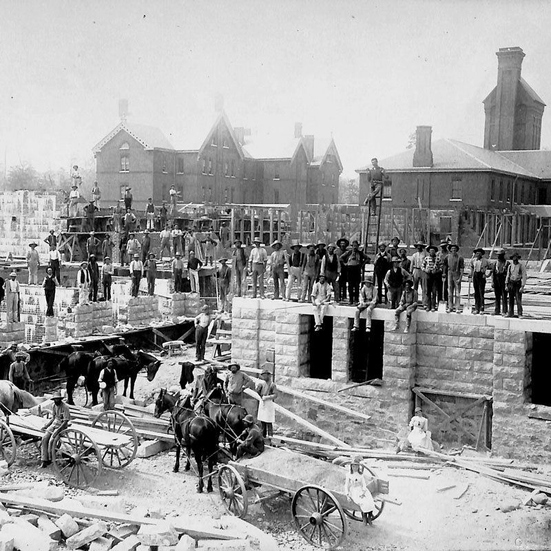 A photograph showing the construction of Cottages A and K and the Administration Building. Workers stand and sit on the building’s stone foundations and three teams of horses and two wagons are standing on the ground below. Behind the foundations, Cottage L, a large three storey brick building, is visible, as is the institution’s original powerhouse and chimney. Image from the Orillia Public Library.