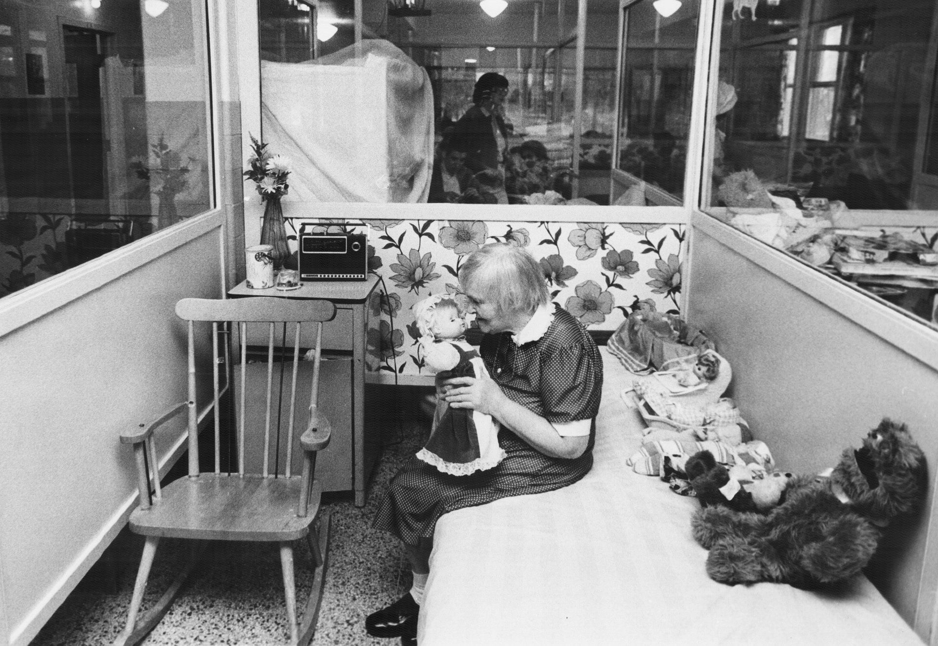Living conditions at the Huronia Regional Centre in 1982. The image shows a small cubicle with walls mostly made out of clear glass. One side of the cubicle is taken up by a narrow bed, on which there is a row of dolls and stuffed toys. Next to this bed is a wooden rocking chair and a small night table with items on top of it, including a vase of flowers and a radio. An elderly woman is sitting on the bed, holding a doll. Given her age, she was likely institutionalized as a child in the early 20th century and spent her entire life in the facility. Image from the Toronto Star Archives.