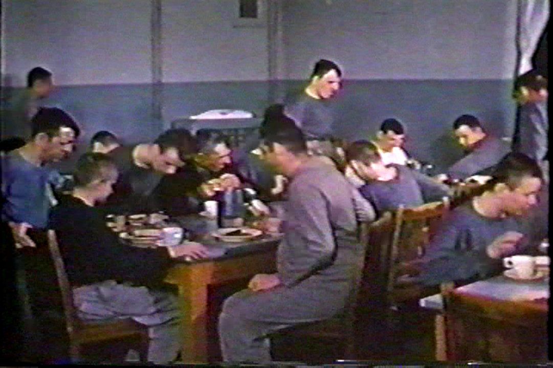 “Custodial” residents eating lunch in a dining room in Cottage D in 1960. Men dressed in institutional clothing are gathered around a table, sitting on worn wooden chairs and eating with plates and cups made of unbreakable melamine. The room has no windows and the walls are painted in two shades of blue, dark blue below and light blue above. Image from the government propaganda film One On Every Street.