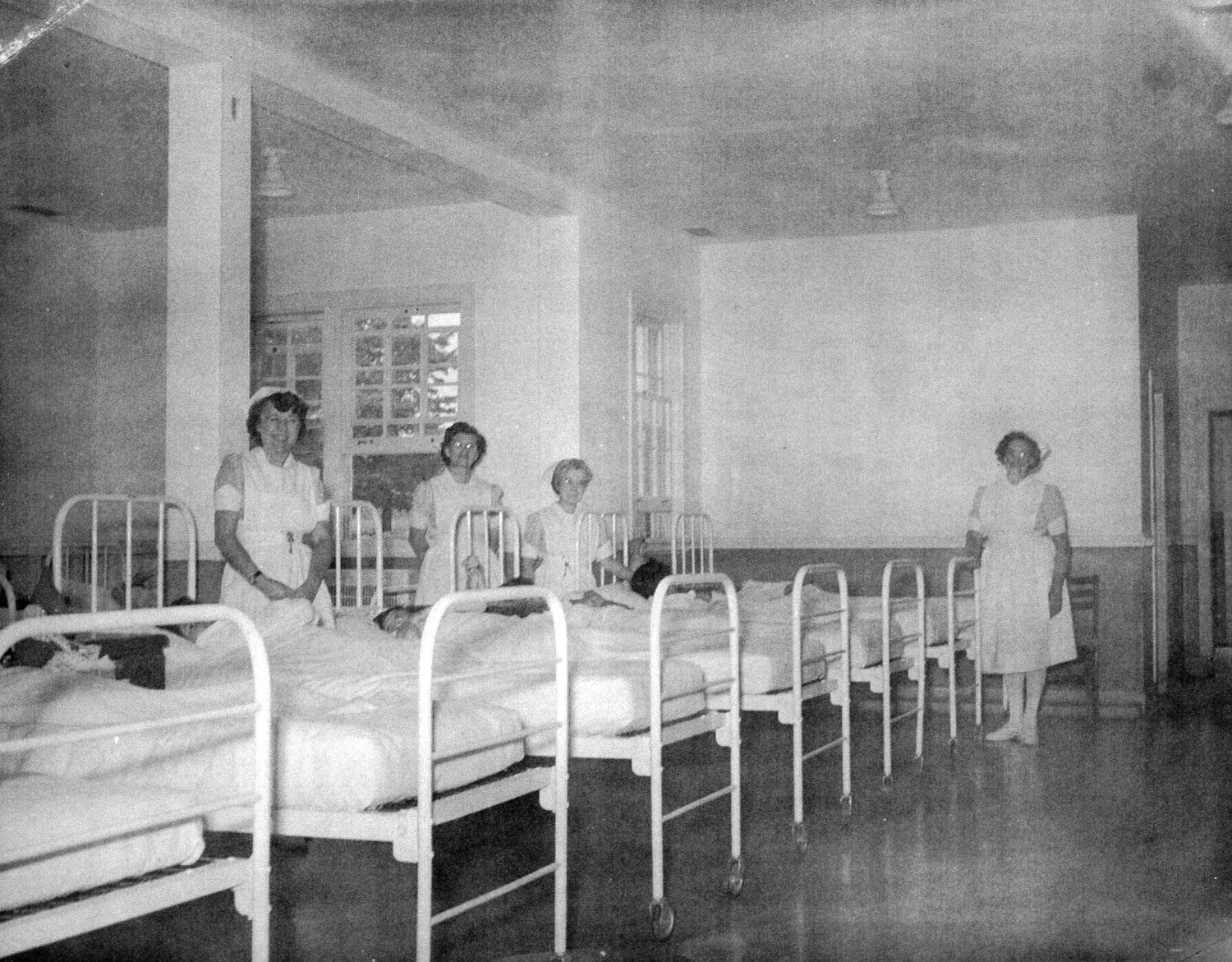Four nurses stand around a row of six iron cots in the newly built infirmary/medical unit of the Ontario Hospital School, Orillia. Children can be seen lying in some of these beds. Other than these cots and a straight backed wooden chair, the room is empty. Many of those who died at the institution died as a result of preventable illnesses. Image from the Regional Centre Records.