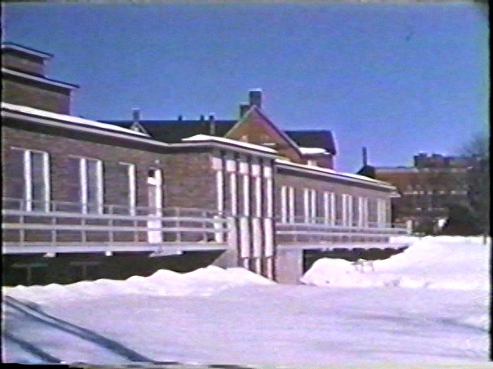 The Huronia Regional Centre’s isolation ward in 1960 in an image from One On Every Street. Beginning in the 1950s, the basement of the isolation ward was the location of the institution’s in house morgue. It is a one storey brick building with a metal walkway running the full length of the structure. This image was taken in the winter and the building is surrounded by snow banks. In the background, the roof of Cottage L is visible, as is the recreation hall/gymnasium. Image from the government propaganda film One On Every Street.
