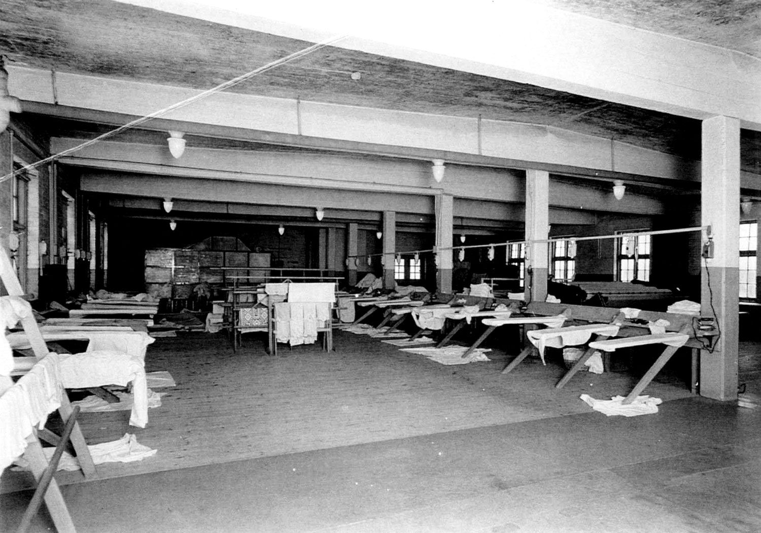 The institution laundry as it would have appeared in the 1930s. It is a large room and appears to be in the basement of one of the institution cottages. Seven support beams run down the centre of the room and two rows of ironing boards occupy its left side, along with two racks with sheets hanging over them. The other side of the room is occupied by several large mangles used for ringing out sheets. Image from the Regional Centre Records.