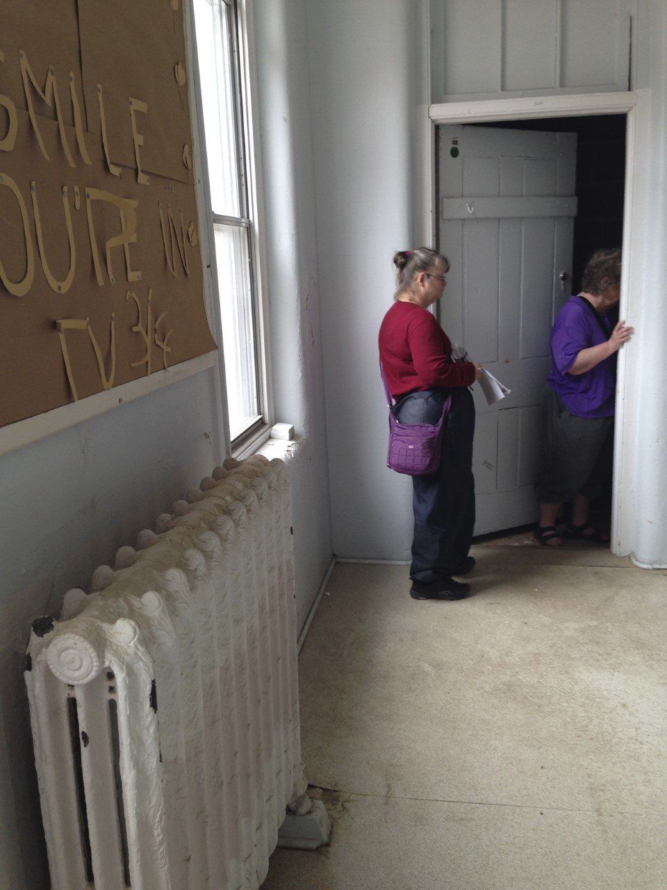 Three images of a pipe room in Cottage K. The first image shows HRC survivors Marie Slark (left) and Pat Seth (right) in an empty dormitory. Pat is standing in the doorway of the pipe room and Marie is standing behind her. In the foreground is an old-fashioned radiator and above that is a bulletin board. Pinned onto the bulletin board are the words “Smile you’re in TV (Terrace View) 3/4.” The second image shows the inside of the pipe room, a narrow space with pipes running along each wall. The left wall is covered in brown tiles, although several of these have been broken off, and the right wall is bare cinderblock. The floor is dirty and covered in garbage. The third image shows the backside of the pipe room door. It is a wooden door painted dark gray and countless deep scratches are dug into its surface.