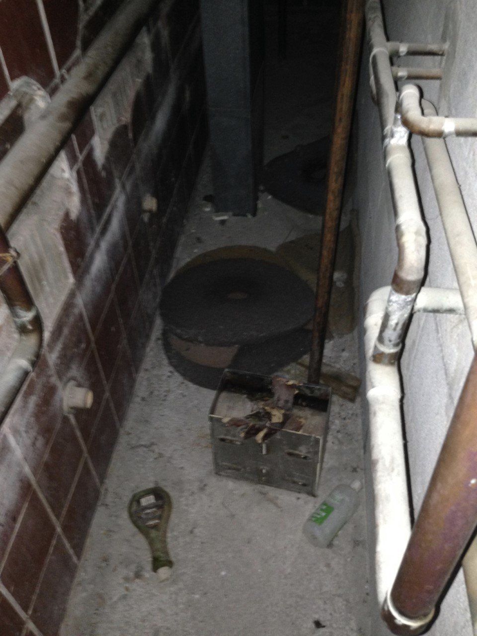 Three images of a pipe room in Cottage K. The first image shows HRC survivors Marie Slark (left) and Pat Seth (right) in an empty dormitory. Pat is standing in the doorway of the pipe room and Marie is standing behind her. In the foreground is an old-fashioned radiator and above that is a bulletin board. Pinned onto the bulletin board are the words “Smile you’re in TV (Terrace View) 3/4.” The second image shows the inside of the pipe room, a narrow space with pipes running along each wall. The left wall is covered in brown tiles, although several of these have been broken off, and the right wall is bare cinderblock. The floor is dirty and covered in garbage. The third image shows the backside of the pipe room door. It is a wooden door painted dark gray and countless deep scratches are dug into its surface.