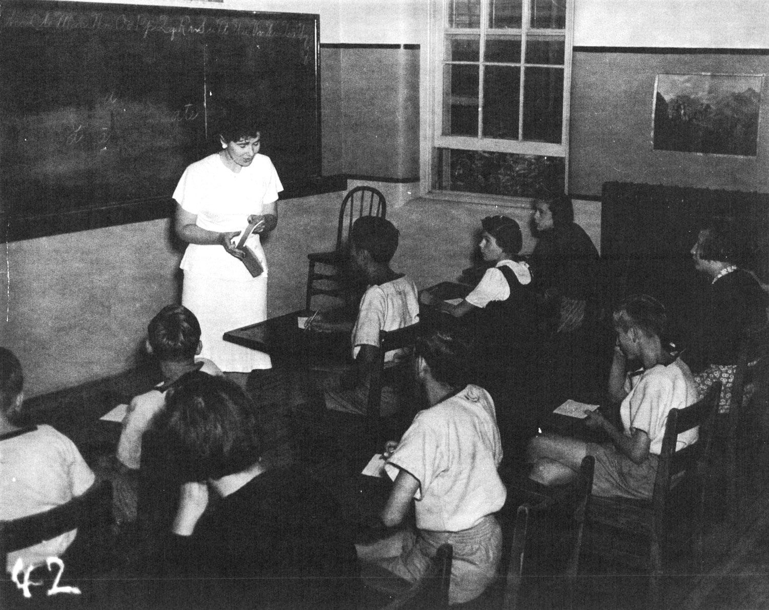 A classroom in the institution school in 1946. Nine children, boys and girls, sit at desks and watch a young female teacher who stands in front of the blackboard holding a book. Very few residents were allowed to attend school and the education they received was extremely substandard. Image from the Regional Centre Records.