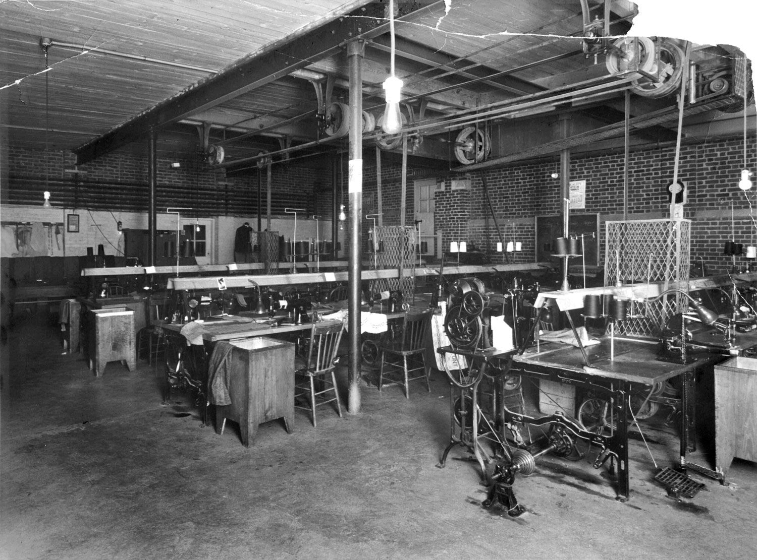 This photograph shows a sewing shop at the Huronia Regional Centre. Three long tables fill the mostly windowless room, with eight industrial sewing machines mounted on each. Three sets of belts and pulleys run along the ceiling and down to the machines. Women and girls incarcerated at the Huronia Regional Centre worked as forced unpaid labour in shops like these, making institutional clothing and straightjackets. Image from the Regional Centre Records.