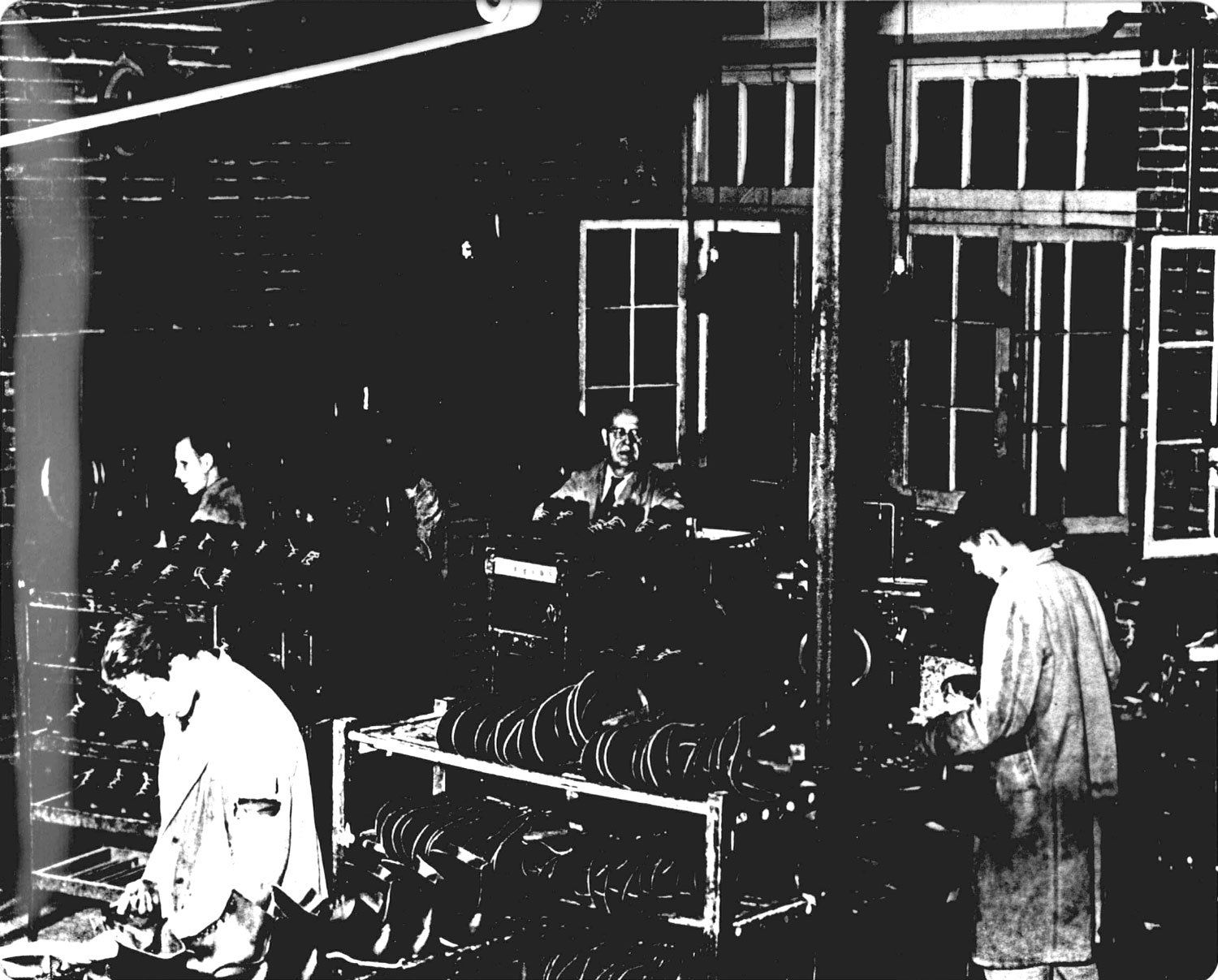 A photograph of the institution shoe shop, where male residents worked making shoes and boots for the Huronia Regional Centre and other facilities. The image is dark, but we can see three residents dressed in loose fitting institutional clothing standing among low shelves filled with boots and shoes. An older male staff member supervises these residents. Belts and pulleys run overhead and one wall is made up mostly of windows. Image from the Regional Centre Records.