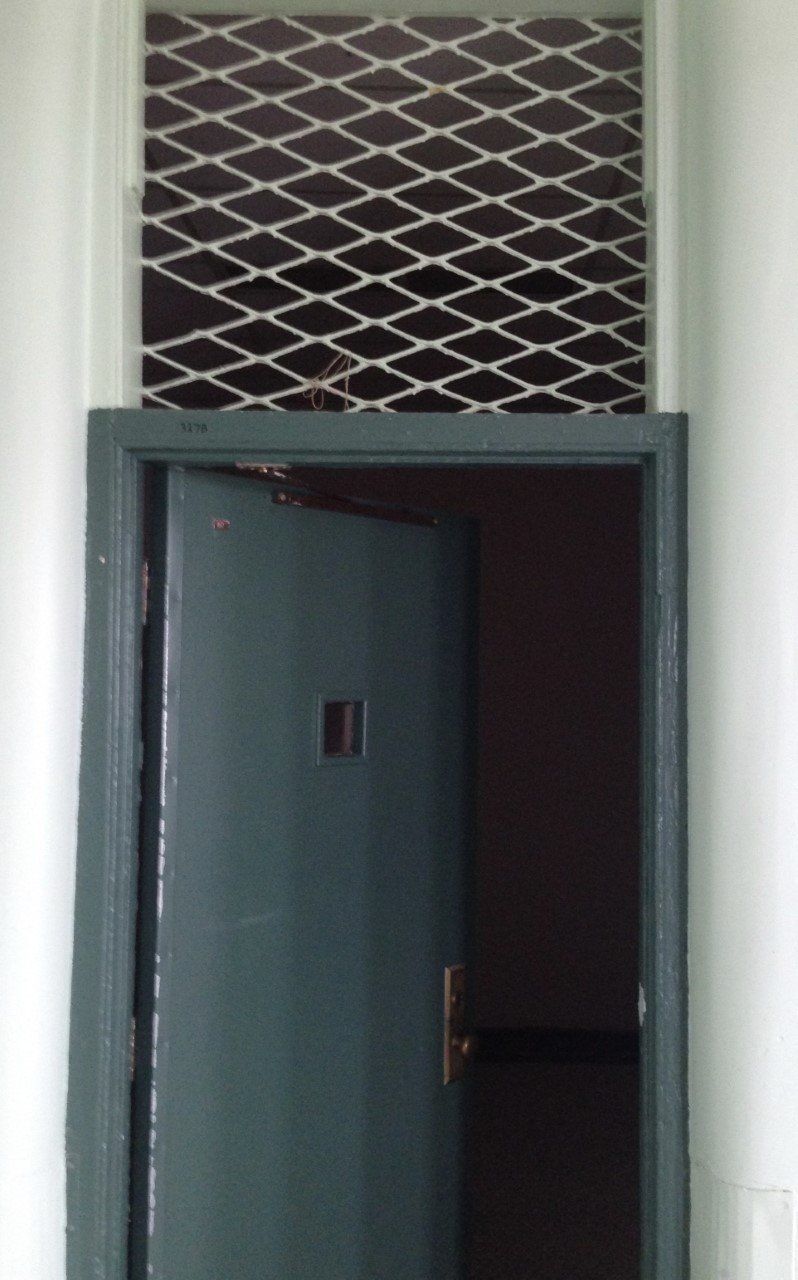 A side/seclusion room in one of the cottages at the Huronia Regional Centre. The image shows a dark green door with a single tiny square window. The door is open, revealing the dark room beyond. Above the door is an opening covered in grating. These openings were added sometime after the 1950s to allow heat from the wards into the previously unheated side rooms. Residents were locked in side rooms for minor infractions with only a mattress (at most) and a bucket to use as a toilet. Evidence suggests that in some cases residents were isolated in side rooms for years at a time