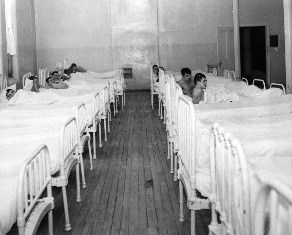 An older ward at the Huronia Regional Centre with wooden floors. Twenty-one iron beds are setup in two rows and at least eleven of these are occupied by naked men. The floor is in poor condition, with boards beginning to separate and at least one hole. Image from the Toronto Star.