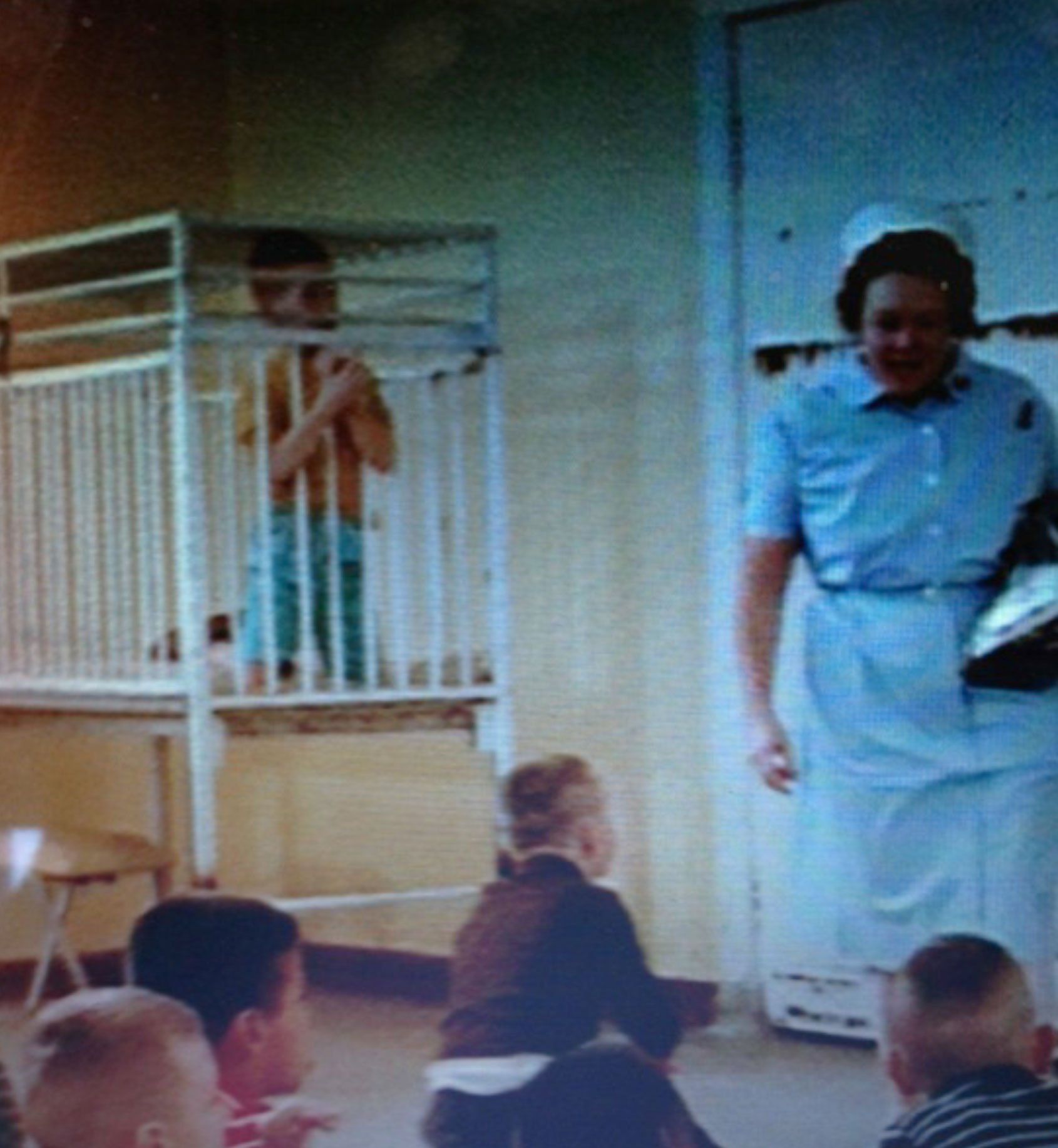 This image shows a children’s dormitory at the Ontario Hospital School, Orillia. Four young boys are sitting on the floor in front of a female staff member wearing a nurse’s uniform. Next to this staff member is a cage cot. A young boy is locked in the cot, sitting up on his knees because he has no room to stand. He has extended his arms through the bars of the cage and his hands are clasped below his chin. Behind the staff member is a huge steel door. Rust bleeds through the door’s white paint. Image from the National Film Board of Canada documentary Danny and Nicky.