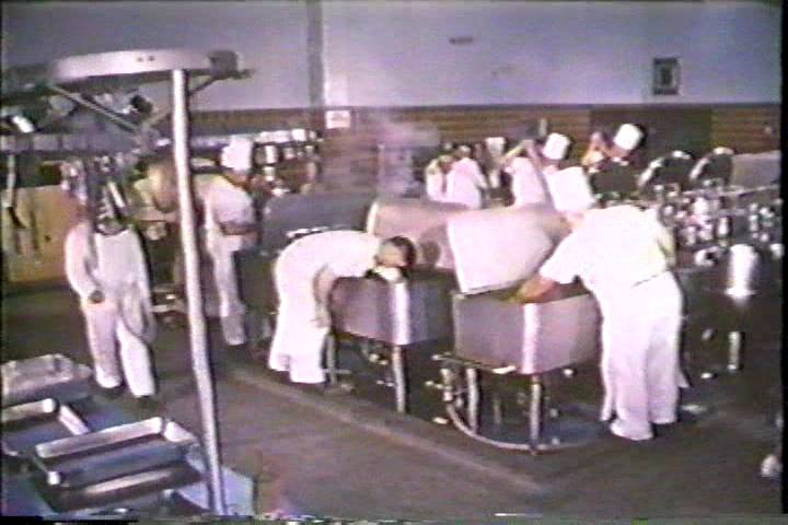 An image of the large central kitchen at the Ontario Hospital School, Orillia. In the foreground, three male staff members wearing white chef’s clothes are bent over large stainless steel tubs filled with food. Another man dressed in chef’s clothes is walking past them and more are visible in the background. The far wall is visible and half of it is covered in brown tiles. The upper half of the wall appears to be blue plaster. Image from the government propaganda film One On Every Street.