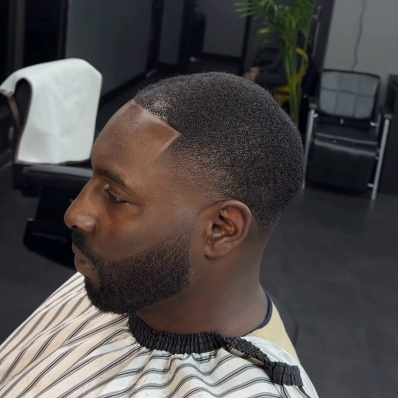 Man with a dark, short haircut and a trimmed beard, wearing a barber's cape in a salon setting.