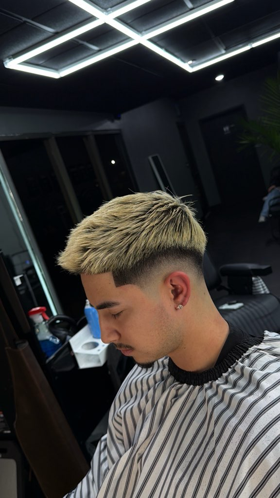 Man with blonde hair getting a haircut. Faded sides, dark eyebrows, striped barber cape. Interior, salon.