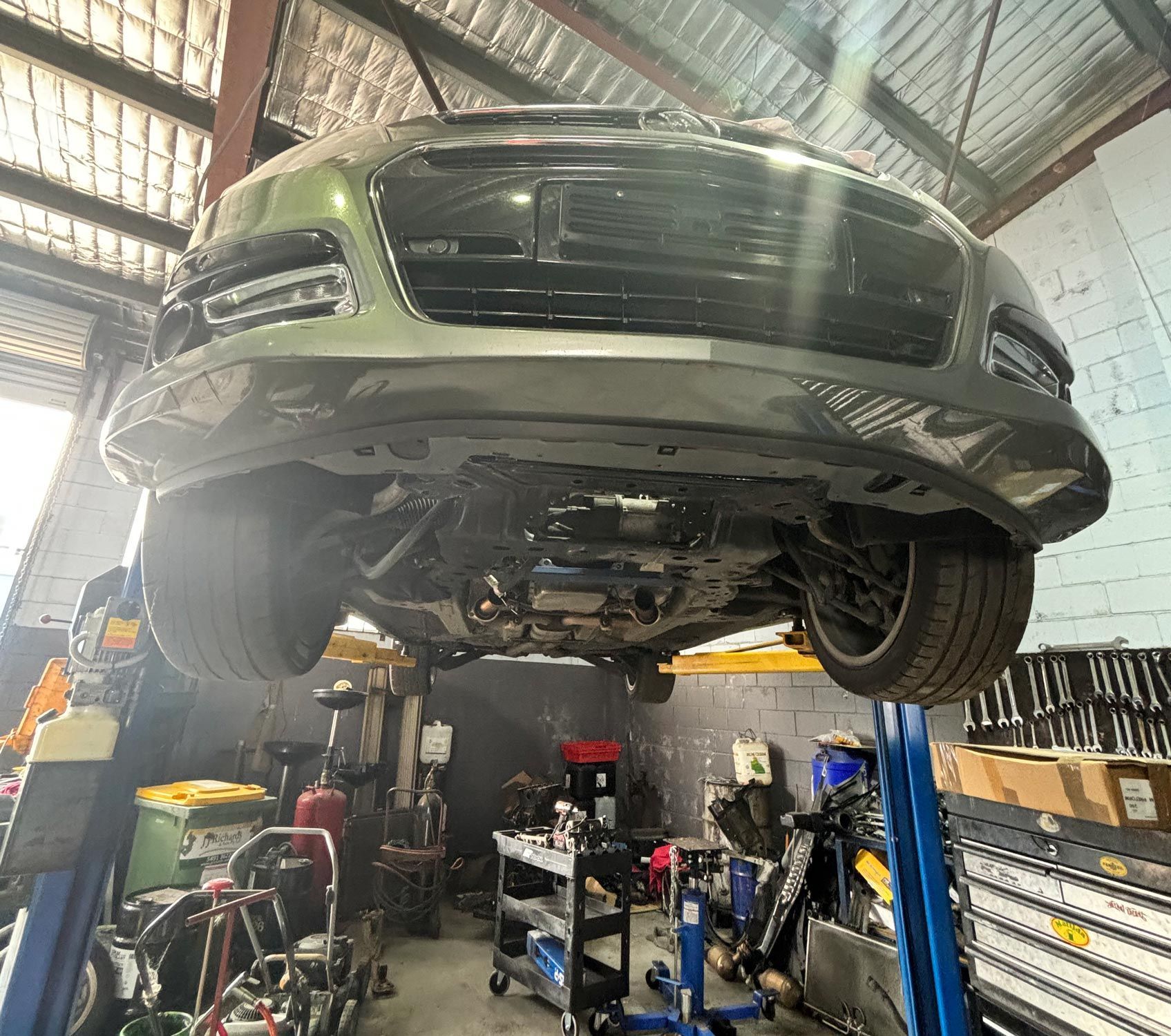 Car raised on a lift in a workshop, viewed from underneath. Gray vehicle; tools and equipment visible.