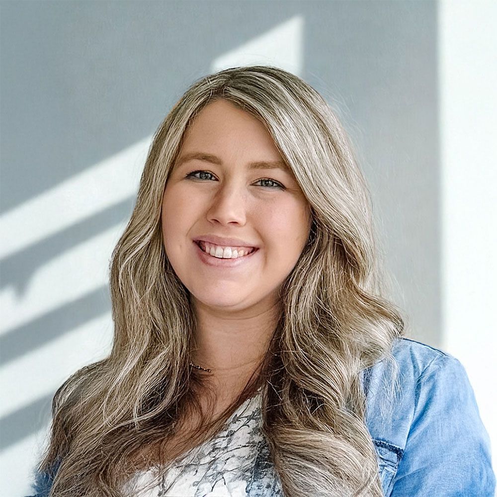 Woman with blonde wavy hair smiles. Wearing a denim jacket and patterned shirt. Lit by sunlight.