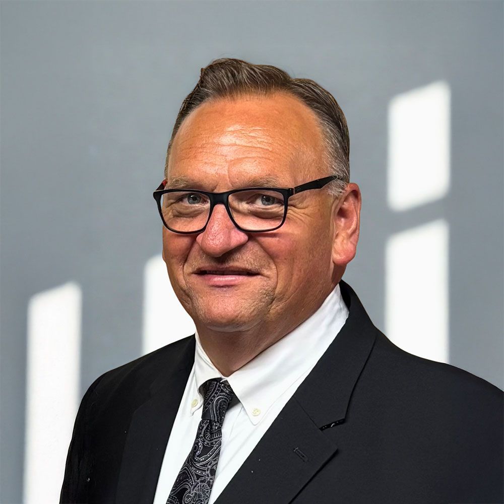 Man in a suit and glasses, smiling, against a gray background.