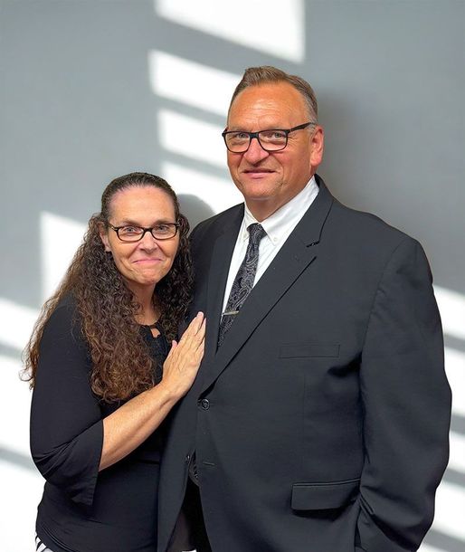 Woman with long brown hair, and man in suit, posing in front of a white wall.