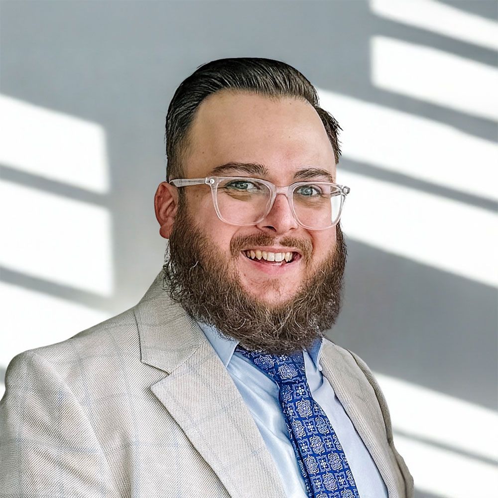 Man with beard and glasses smiles, wearing a light suit jacket and patterned blue tie.