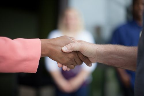 Handshake between a person with darker skin and a person with lighter skin. Blurred figures in background.