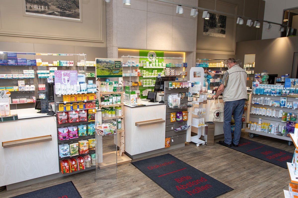 Innenansicht einer Apotheke mit einem Kunden, verschiedenen Medikamenten in Regalen und einer Kasse.