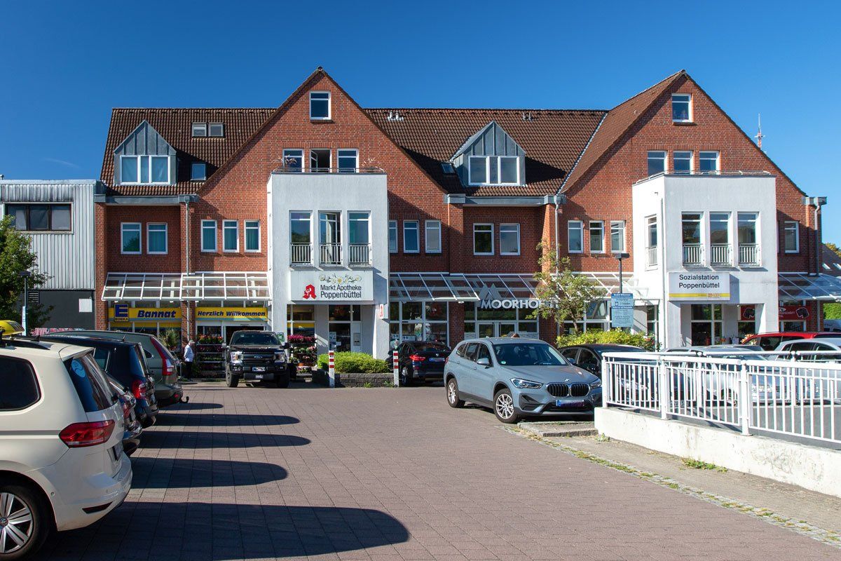 Gebäude mit Geschäften im Erdgeschoss, rote Backsteinfassade mit weißen Akzenten, davor geparkte Autos.