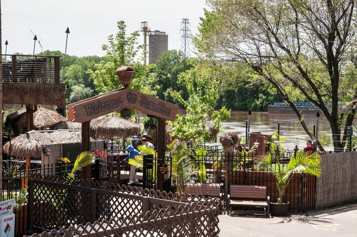 Tropical-themed outdoor restaurant with thatched roofs, plants, and a person near a counter; a river and industrial structures are in the background.