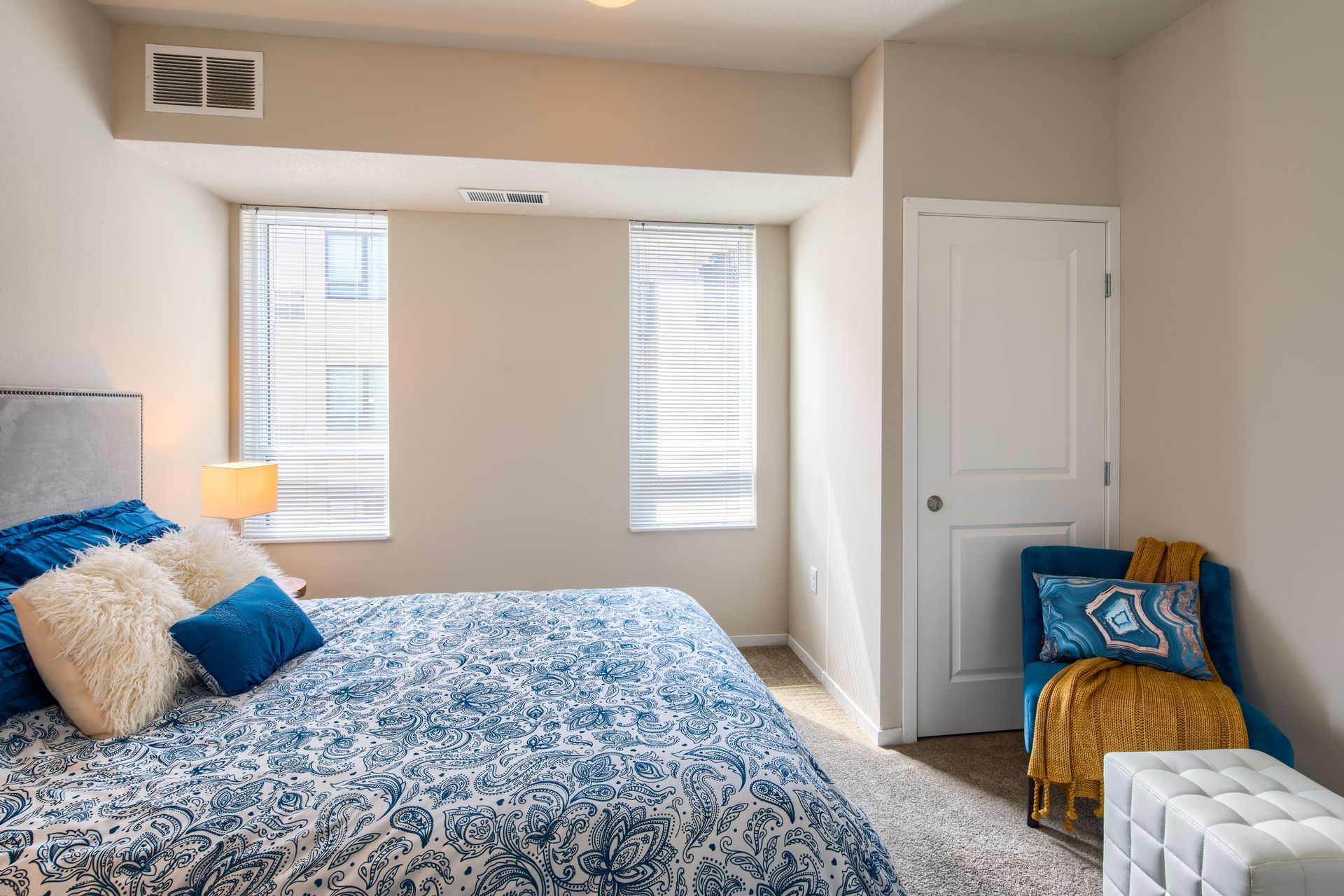 Bedroom with blue patterned bedspread, two windows with blinds, a blue chair with a yellow blanket, and a white door.