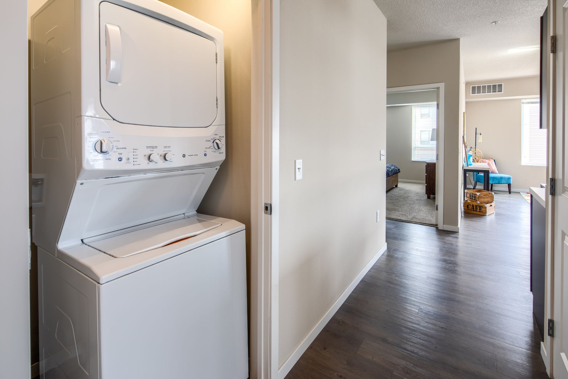 Stacked white washer and dryer in a hallway, view toward a living area with natural light and dark wood floors.