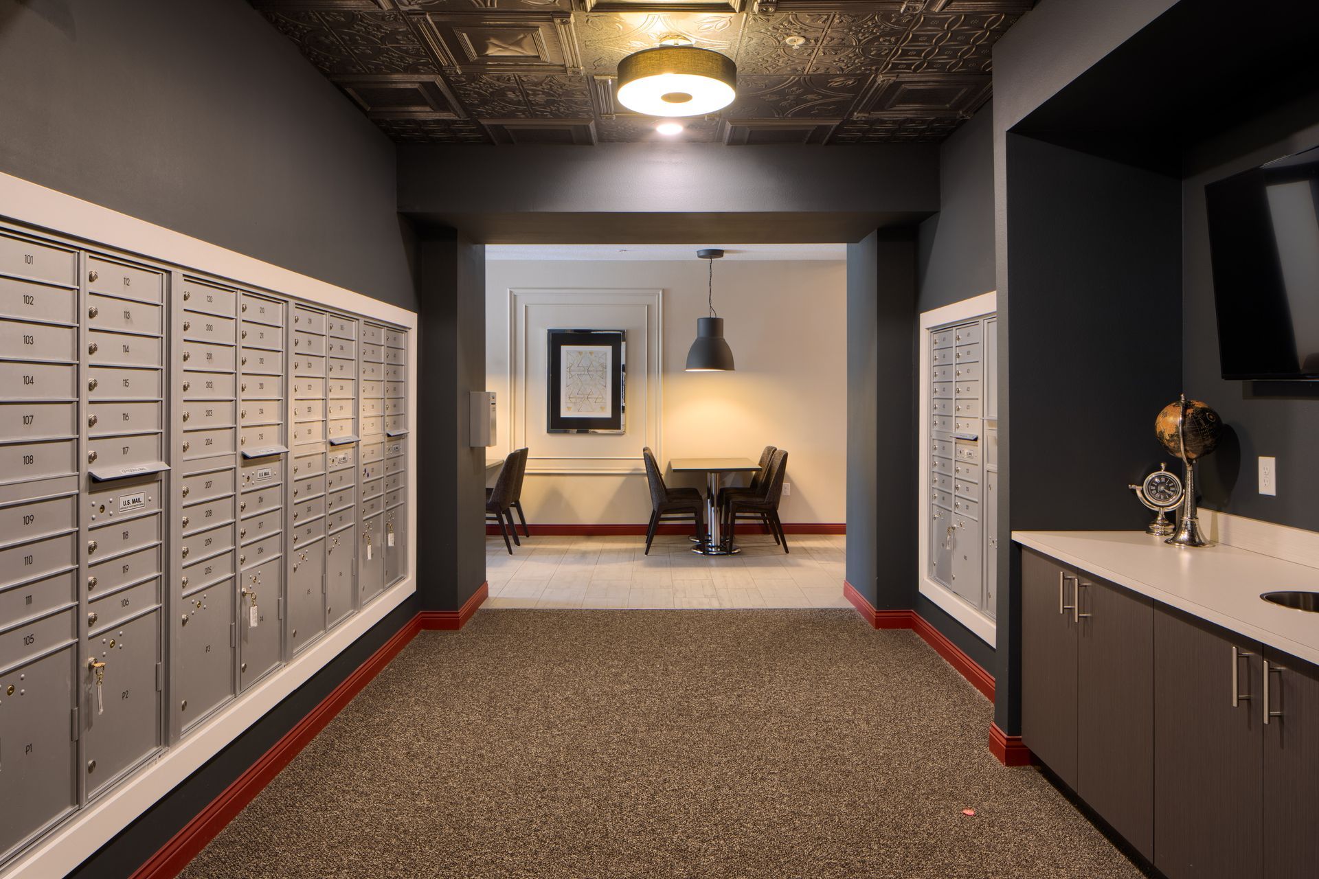 Hallway with mailboxes, a seating area, and a television. Gray walls, patterned ceiling, and speckled carpet.