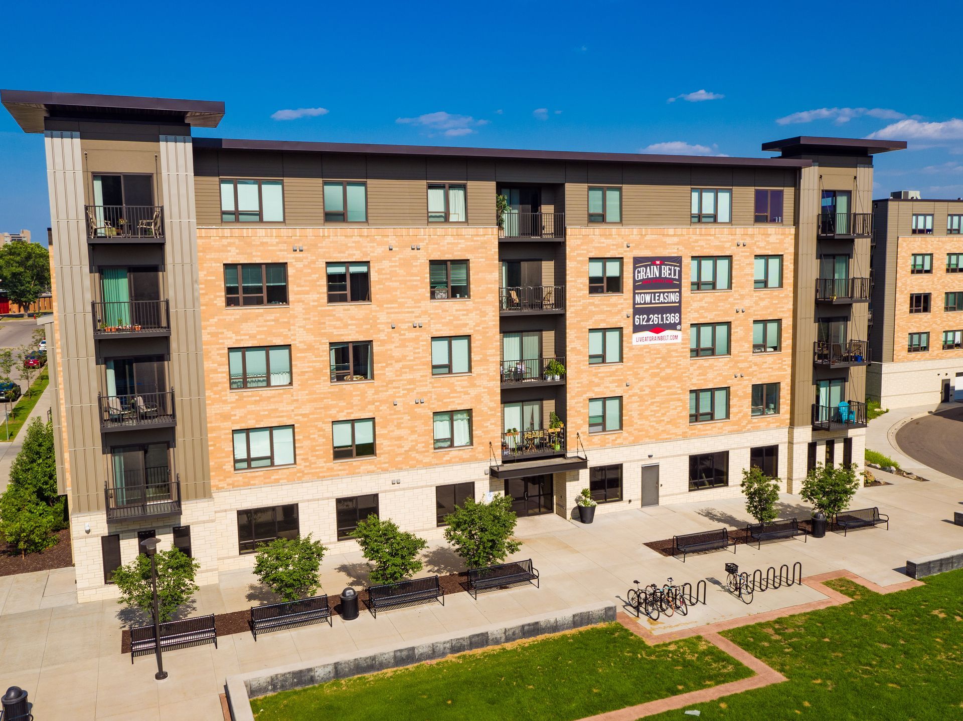 Multi-story brick apartment building with balconies, trees, and green lawn.