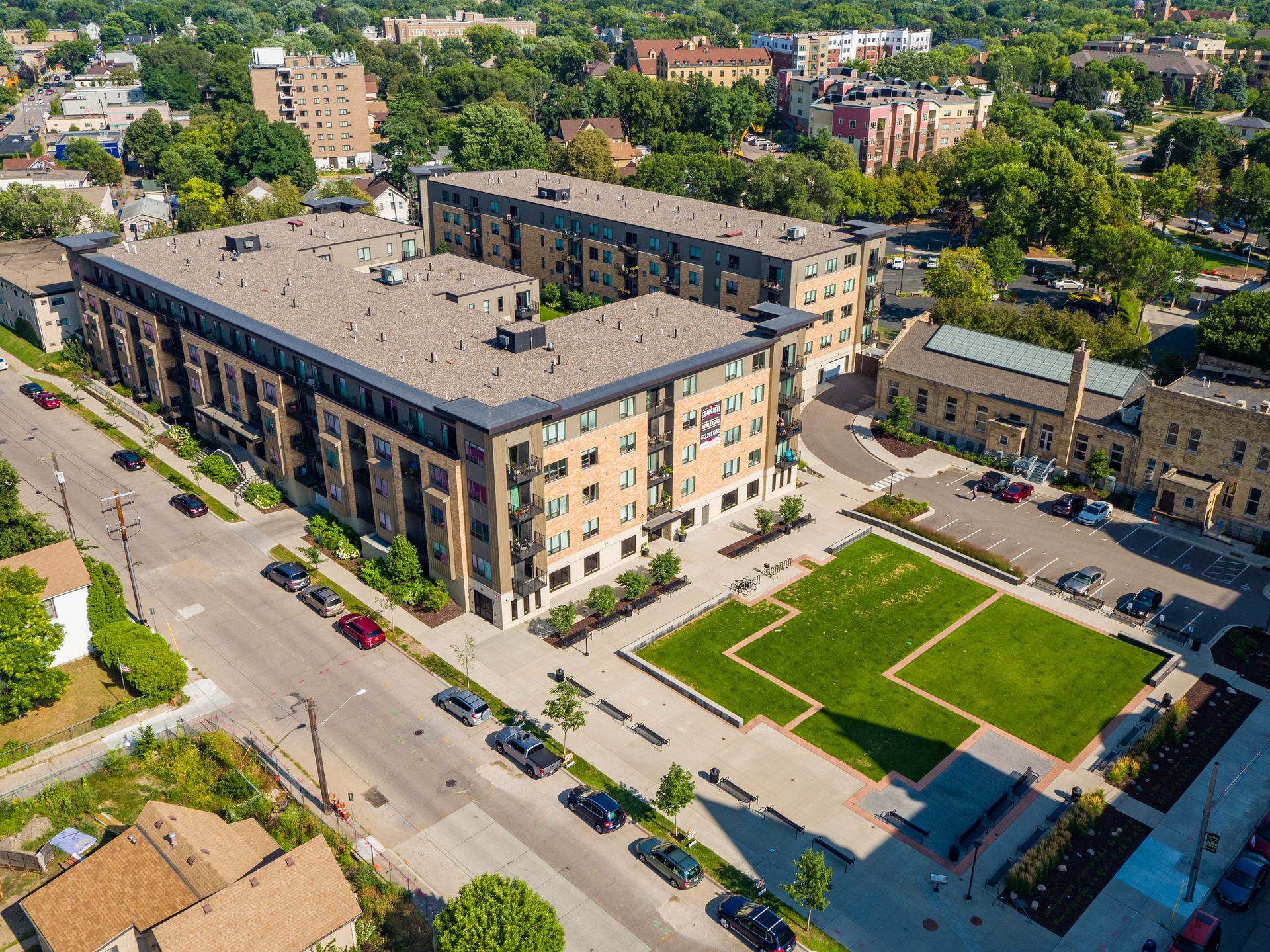 Aerial view of an apartment complex with a green square, trees, and parked cars on a sunny day.
