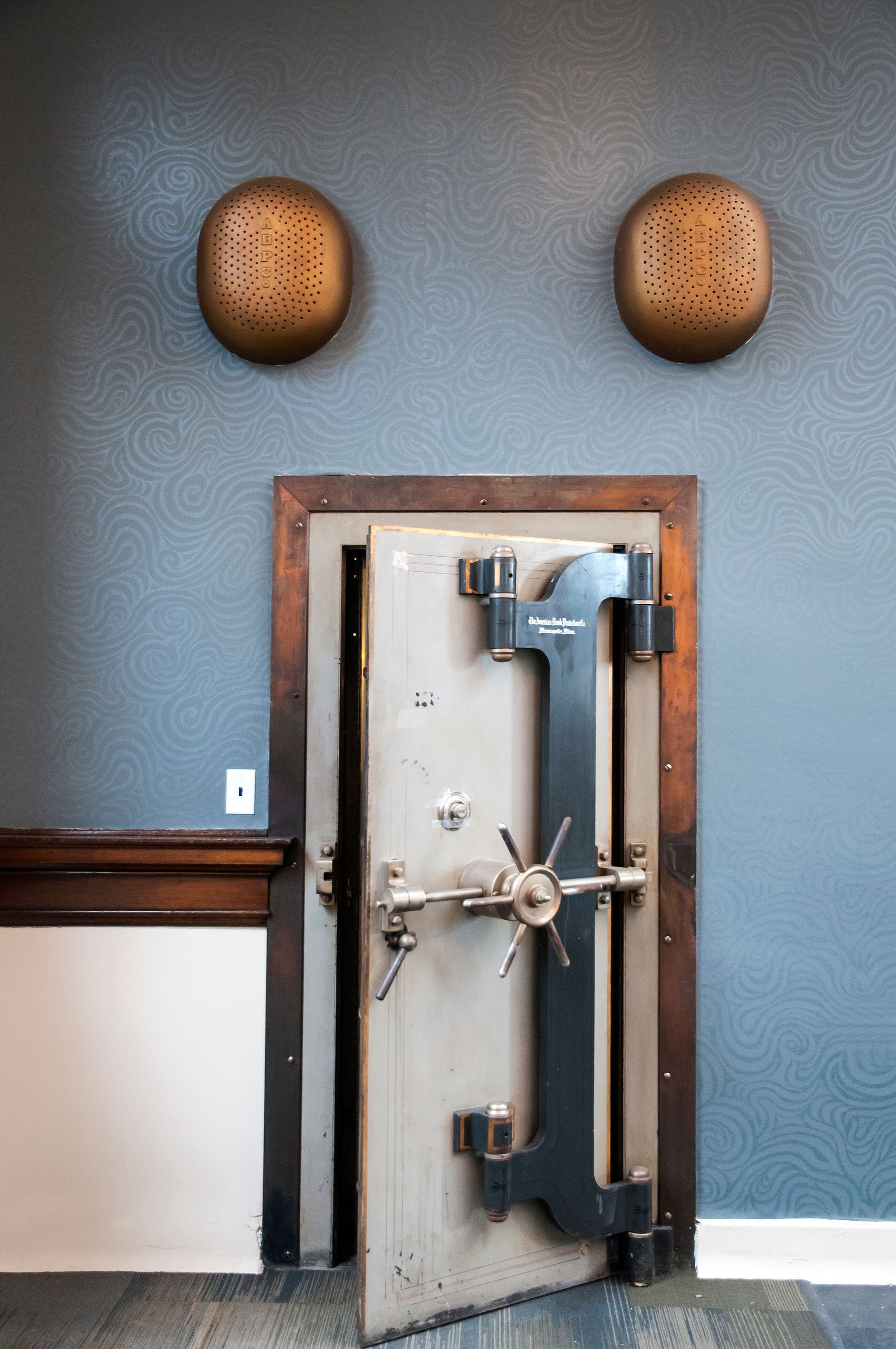 Old bank vault door in a dark blue room, flanked by two circular, perforated wooden decorations.