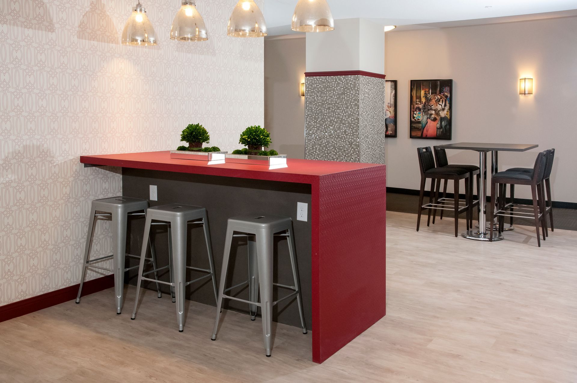 A red bar with silver stools in a modern space, near a textured wall, small plants, and hanging lights.