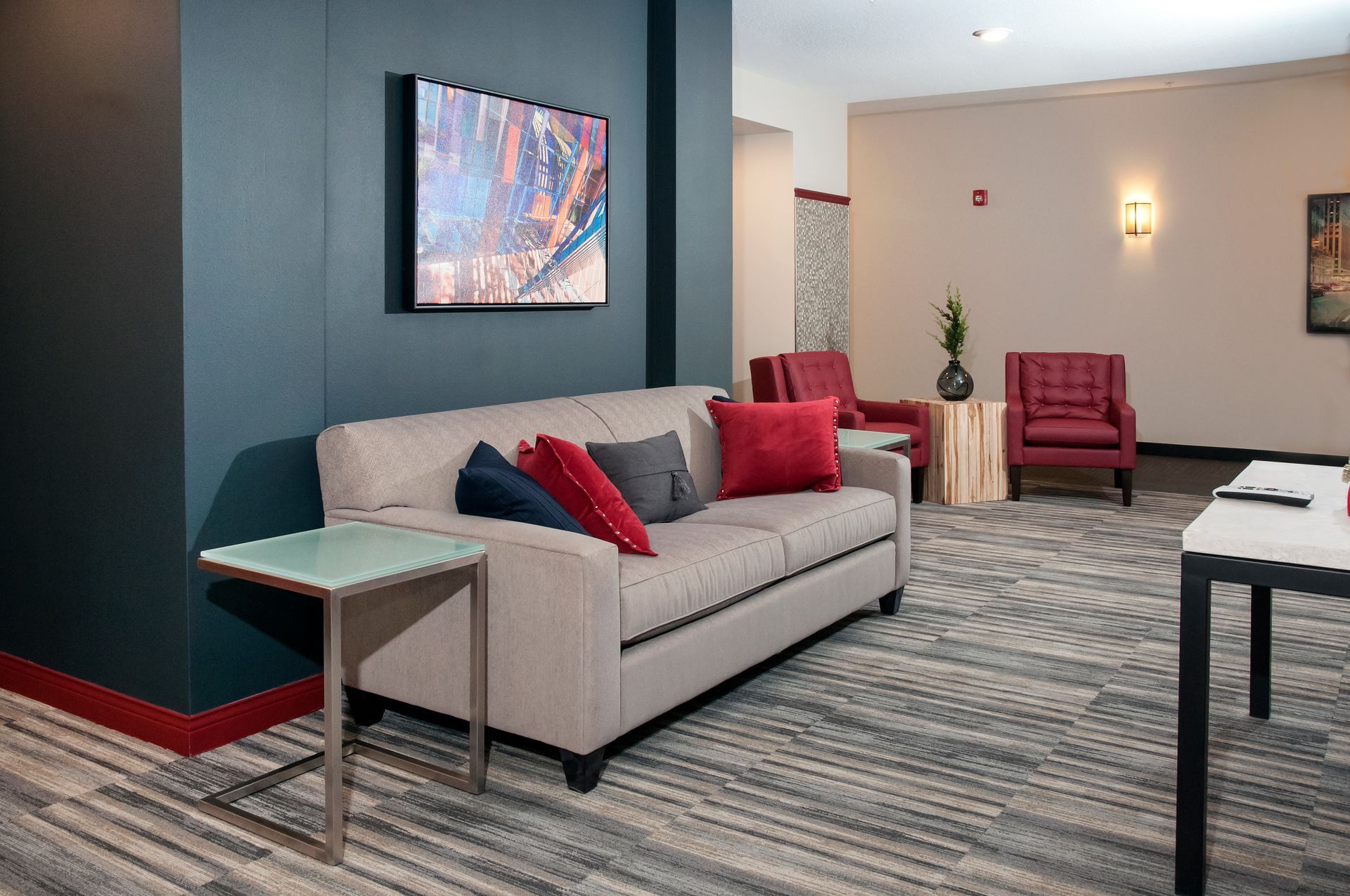 Lounge area with a gray couch and red pillows against a teal wall. Small table, artwork, and chairs visible.