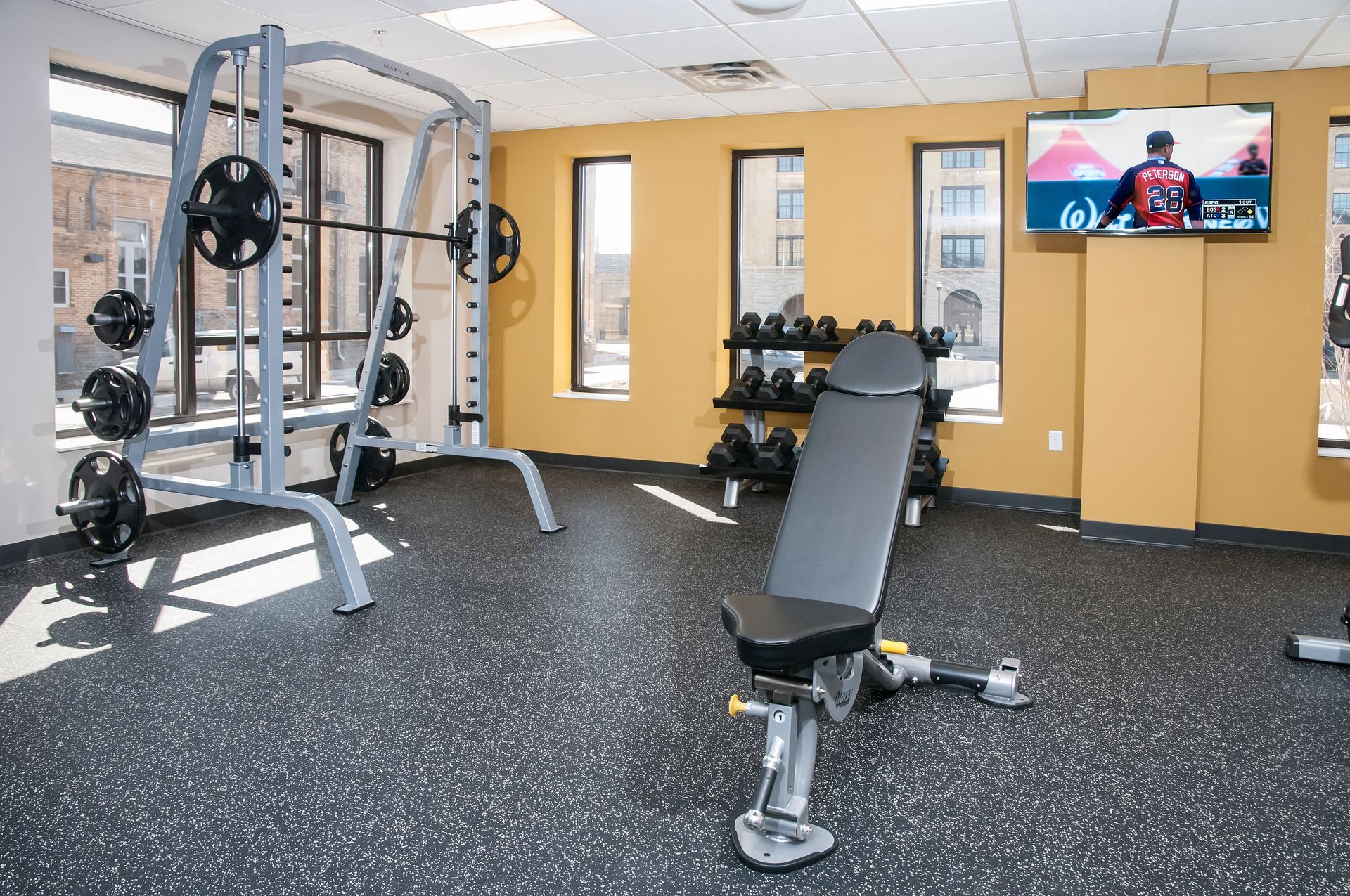 Gym interior: Smith machine, adjustable bench, dumbbells, and TV against mustard-colored walls with windows.