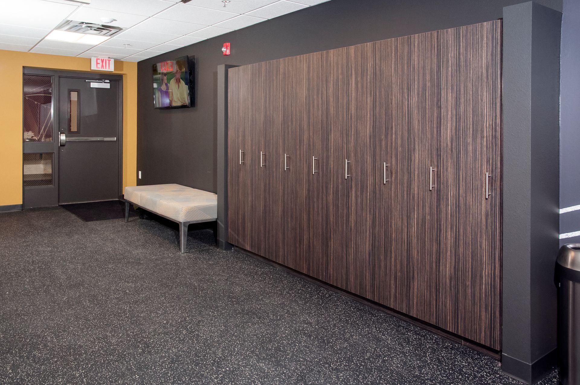 A hallway with dark wood lockers, a bench, a door, and a television. Black and gray speckled floor.