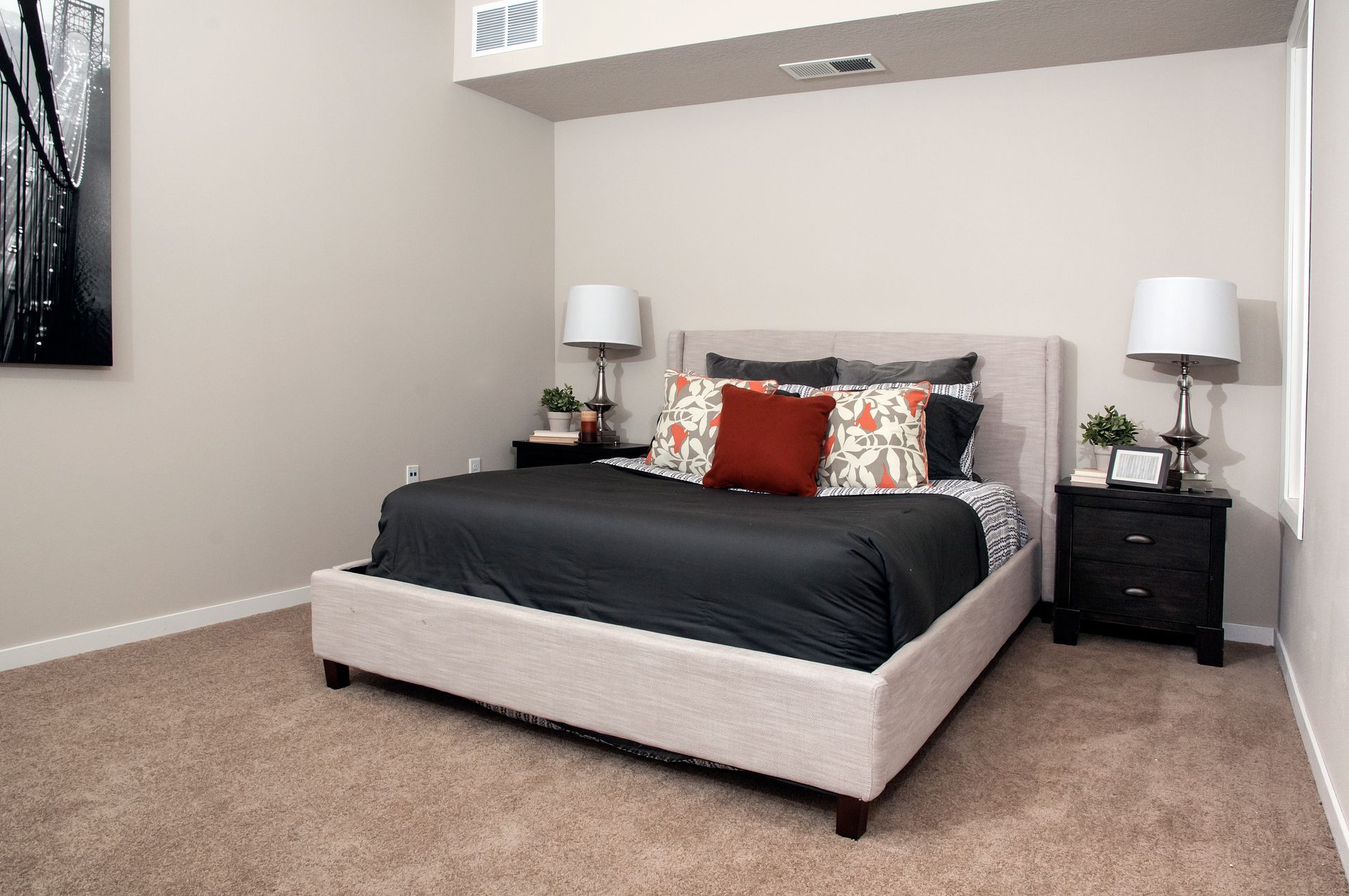 Bedroom with beige walls, bed with dark bedding and decorative pillows, two bedside tables with lamps, and carpet.