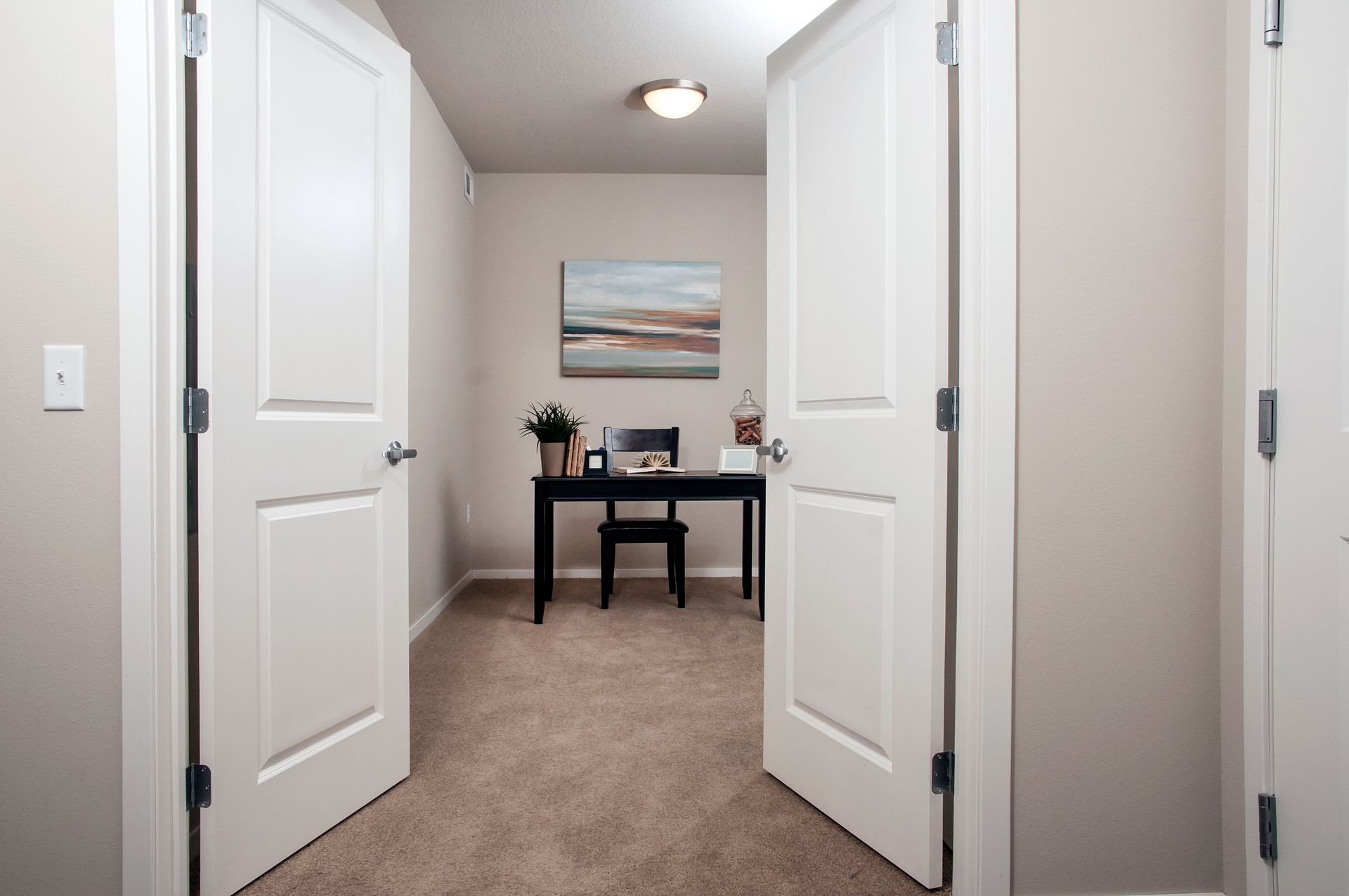 Two white doors open to a small home office with a desk, chair, and artwork.