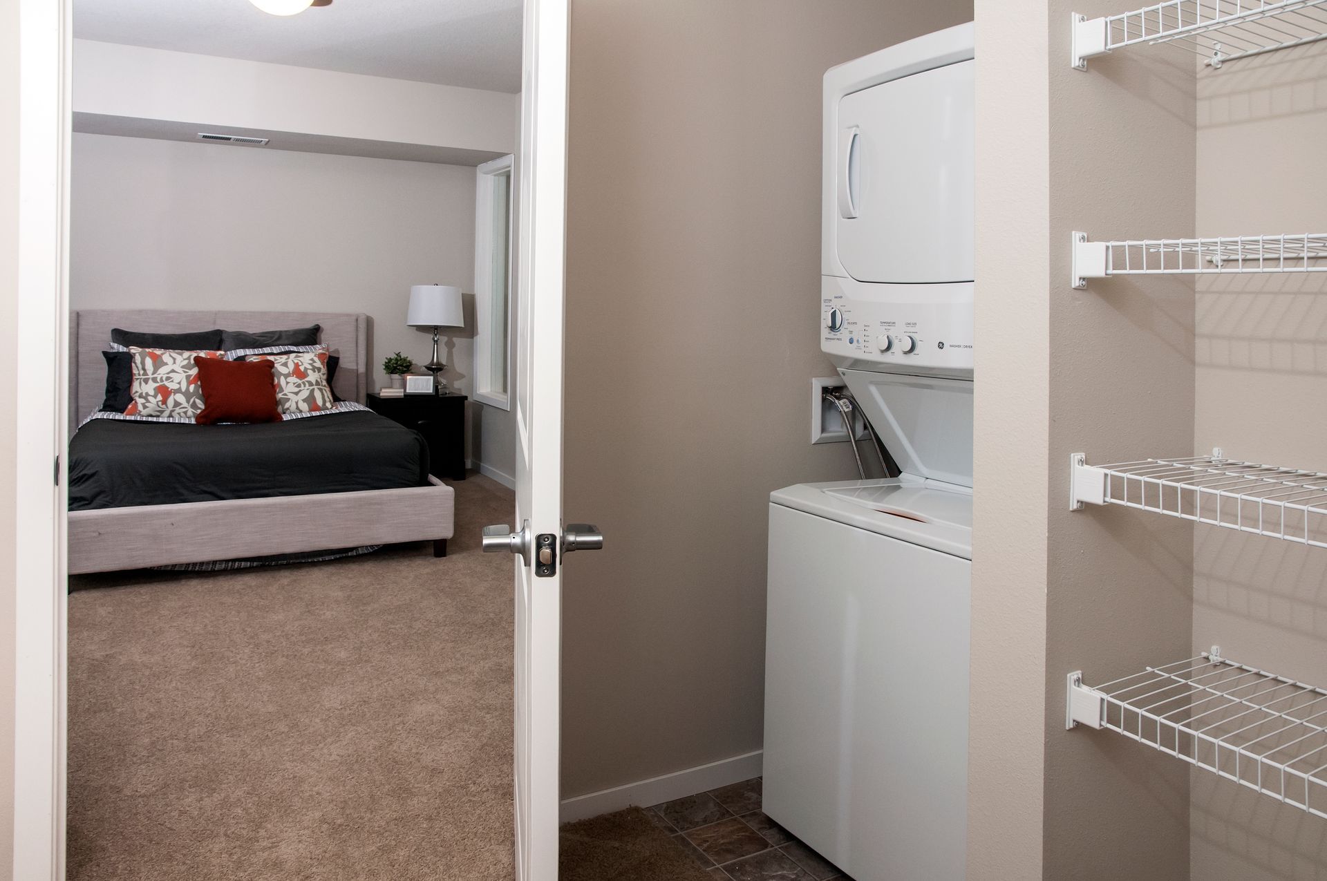 Open door reveals bedroom and stacked washer/dryer in a neutral-toned apartment. Wire shelving is visible.
